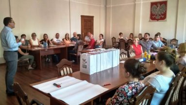 Zdjęcie przedstawia mężczyznę przemawiającego do grupy osób, którzy siedzą przy stołach