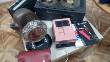 Na stoliku przedmioty z czasów PRL-u adapter, projektor, magnetofon kasetowy, płyta wideo, zegar, aparat do zdjęć