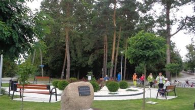 Grupa osób w parku dużo zieleni drzewa, krzewy, ławki, kamień pamiątkowy z tablicą upamiętniającą ofiary wojny
