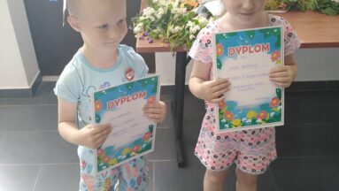 Dzieci z wiankami na głowie pozują do zdjęcia, pokazując swoje dyplomy