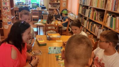 Zdjęcie przedstawia kobietę z dziećmi. Wszyscy siedzą przy stolikach i grają w gry planszowe. W tle regały z książkami