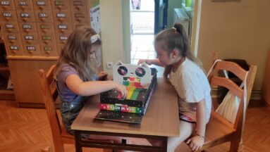 Zdjęcie przedstawia dziewczynki grające w gry planszowe. W tle regały z książkami