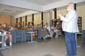 Mężczyzna stoi i opowiada o czymś. Na sali przy stolikach siedzą inni ludzi i słuchają jego opowieści