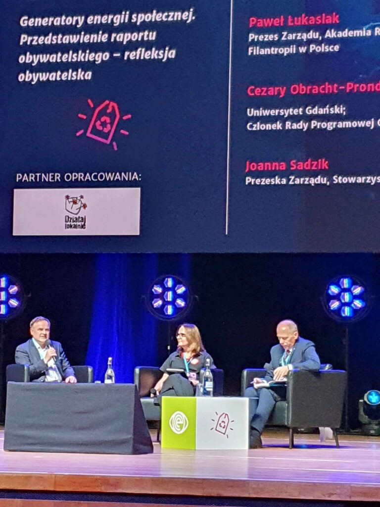 Panel dyskusyjny, 3 osoby opowiadają o tworzeniu raportu obywatelskiego. Mikrofon trzyma Paweł Łukasiak. W tle na wyświetlaczu nazwiska os&oacute;b biorących udział w dyskusji 