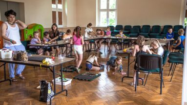 Na dużej sali, prawdopodobnie w szkole, przy stolikach siedzą dzieci. Są z nimi dorośli, którzy coś tłumaczom. Na podłodze jak i na stolikach znajdują się przedmioty w stylu ekologicznym, być może z recyklingu