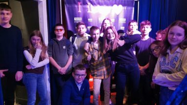 Na fotografii znajduje się grupa osób, które pozują w teatrze. Światło w pomieszczeniu jest przyciemnione, niebieskie. W tle, za grupą znajduje się granatowa kurtyna