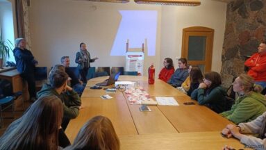 W dużej sali przy podłużnym stole siedzą młodzi ludzie. Na końcu sali dorosła osoba opowiada coś przy prezentacji, jaka jest wyświetlana na ścianie