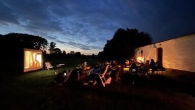 Późne godziny wieczorne, grupa osób siedzi na leżakach na zewnątrz i ogląda film wyświetlany na projektorze.
