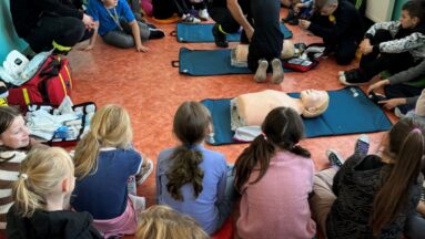 Zdjęcie przedstawia szkolenie z udzielania pierwszej pomocy przy użyciu sprzętu AED prowadzone przez strażaków dla uczniów. W zajęciach uczestniczy duża grupa dzieci.