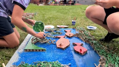 Zdjęcie przedstawia dwie osoby malujące akwarium wykonane z tektury i innych odpadów takich jak folia bombelkowa. W tle widać innych uczestników "Festiwalu kolorów".