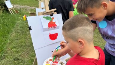 Zdjęcie zrobione podczas pleneru malarskiego w ramach wydarzenia "Festiwal kolorów" w projekcie "Bezpieczna Strefa Chillout". Na pierwszym planie widać dwóch chłopców, którzy nabierają farbę z palety. Przed nimi stoi sztaluga na której namalowano fragment jabłka. W tle widać leżaki i innych uczestników imprezy zgromadzonych na terenie zielonym przy Szkole Podstawowej w Wyczechach.