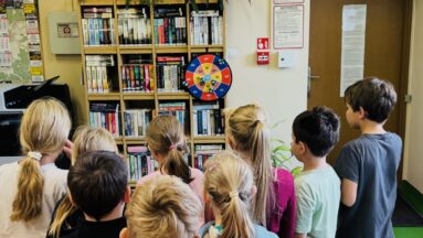 Grupa dzieci stoi tyłem do aparatu i rzuca rzutkami do kolorowej tarczy zawieszonej na bibliotecznym regale.