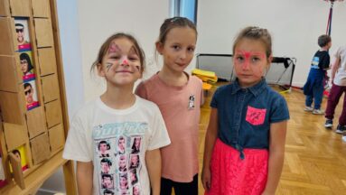 Trzy małe dziewczynki stoją obok siebie w jasnym pokoju i uśmiechają się do aparatu. Mają pomalowane twarze w kolorowe wzory i motyle.