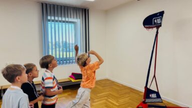 Czwórka chłopców stoi w rządku i obserwuje piłkę lecącą w stronę przenośnej koszykówki z logo NBA.